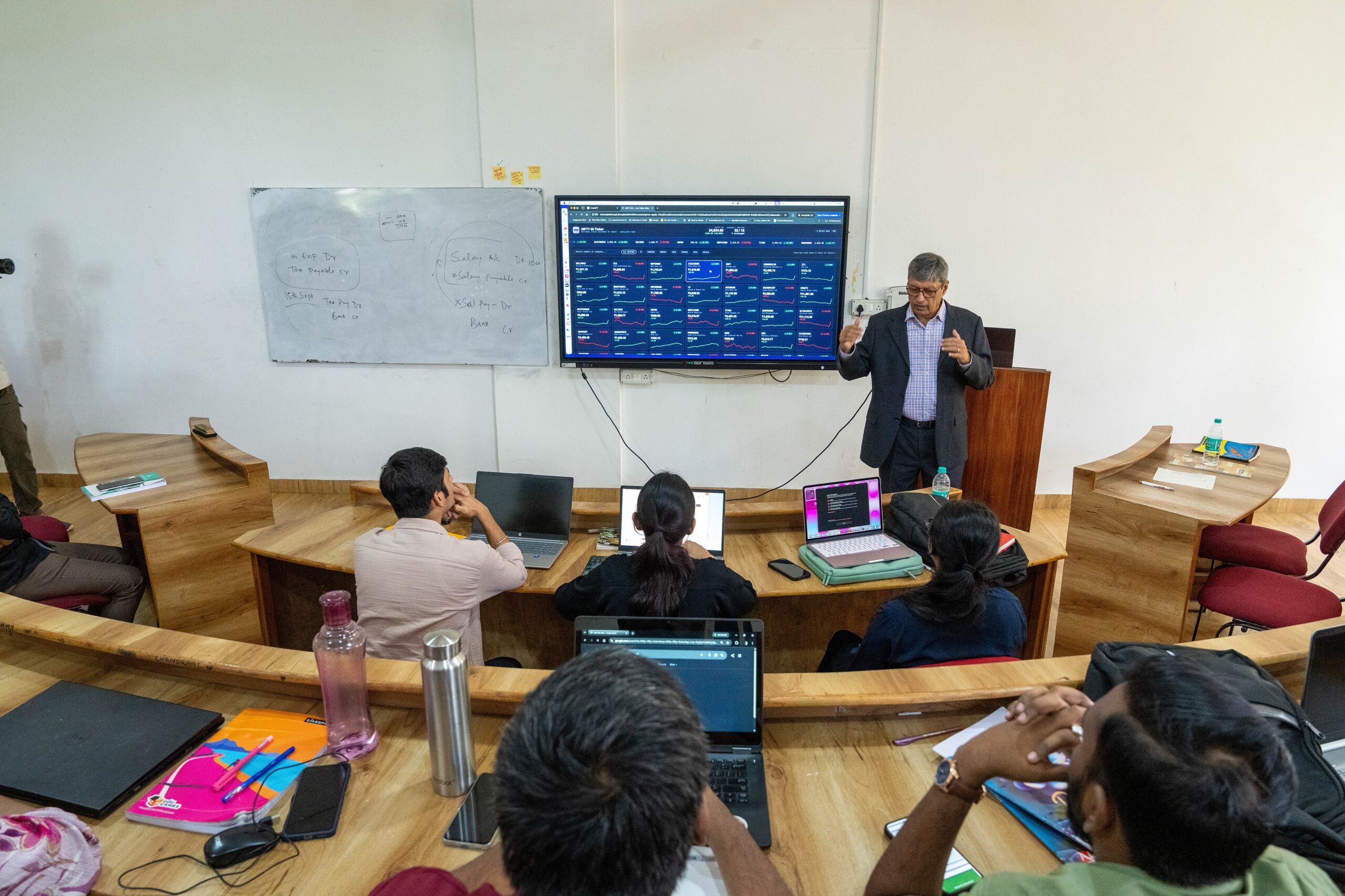 Faculty member teaching with a financial dashboard while students use laptops