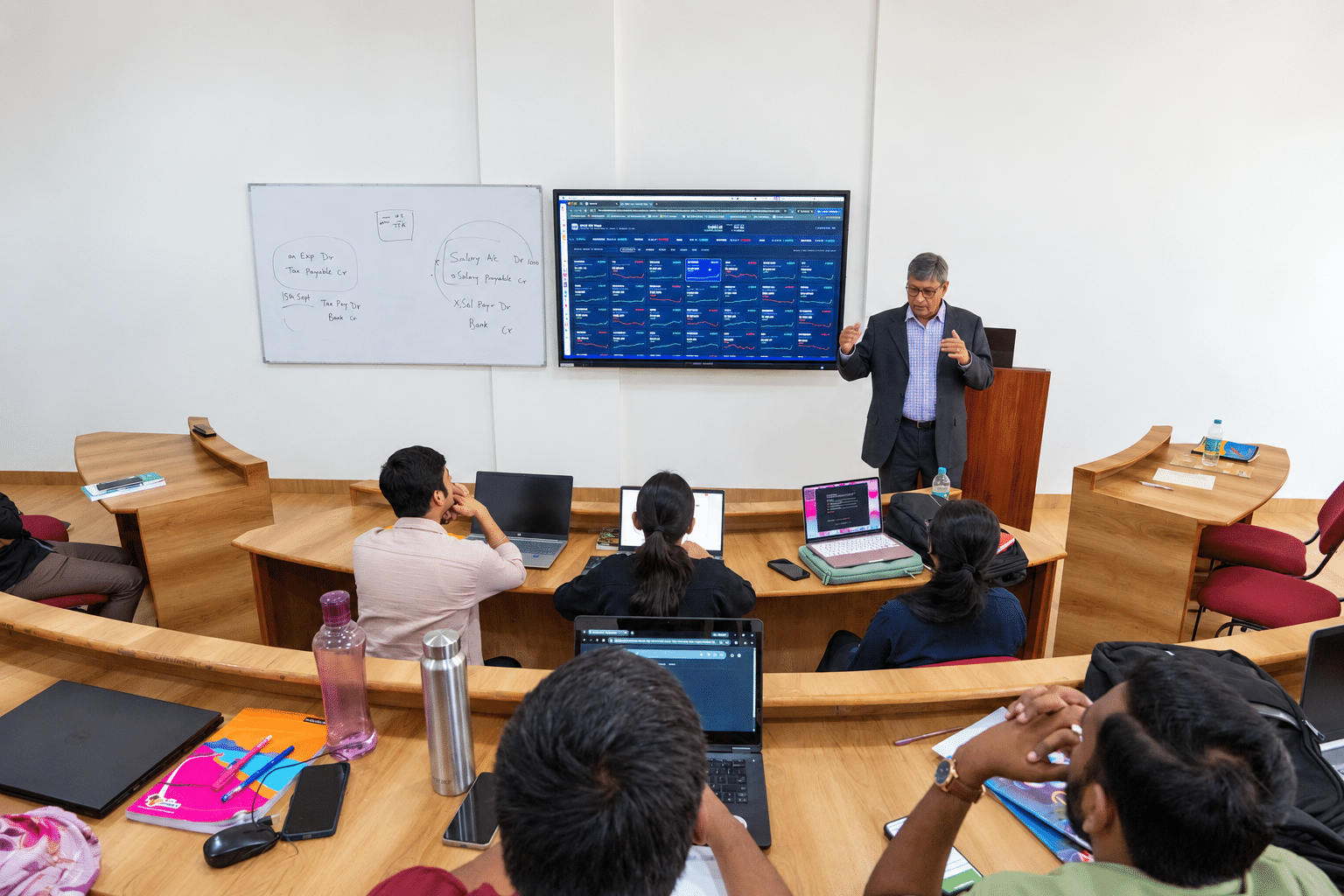 Faculty member teaching with a financial dashboard while students use laptops